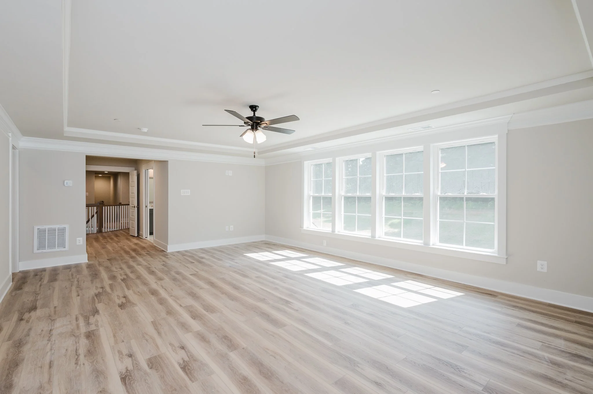 Empty living room with large windows, light wood flooring, beige walls, a ceiling fan, and an opening leading to a hallway.