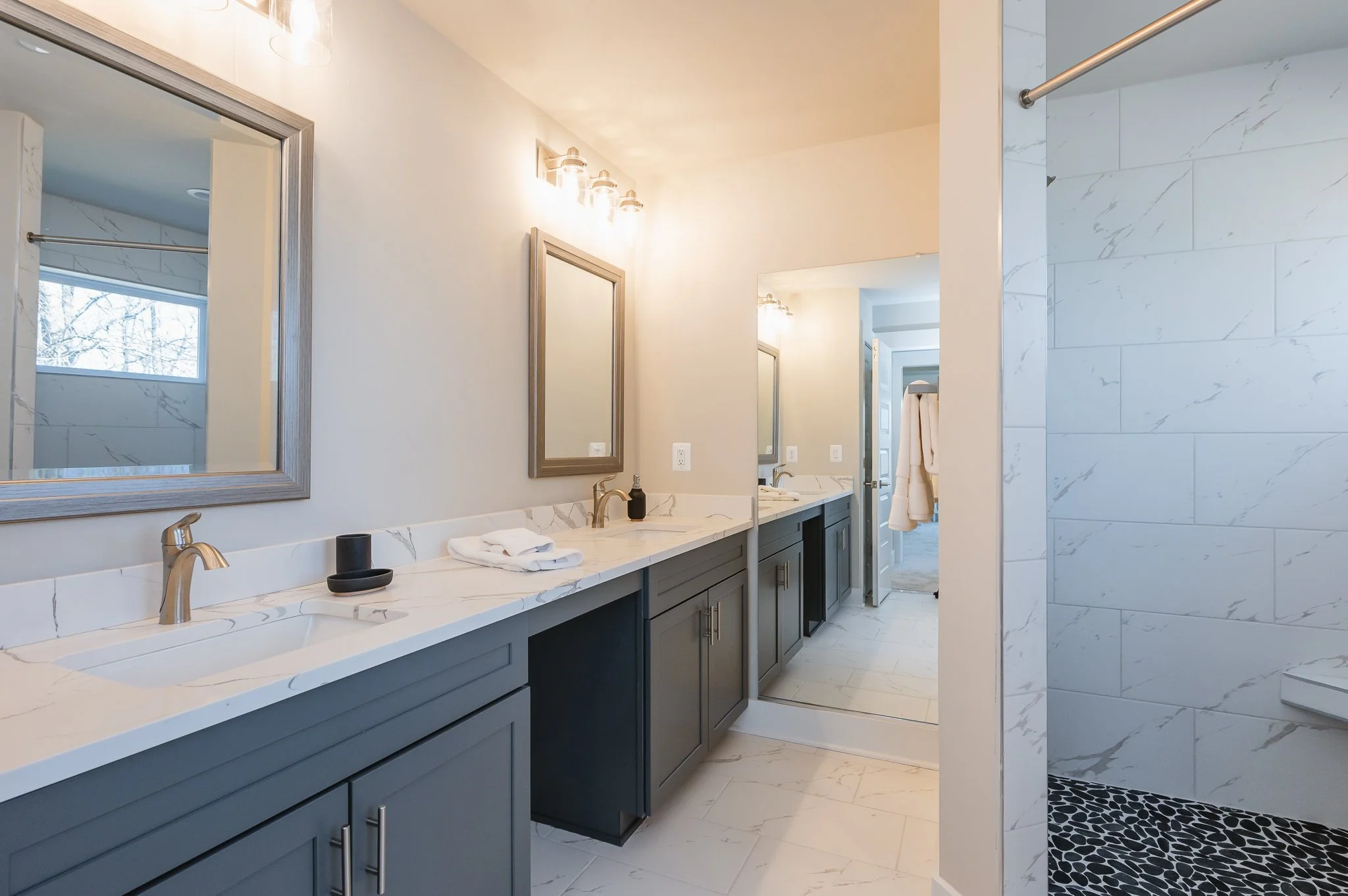 Modern bathroom with dual sinks and large mirrors, light-colored countertops, gray cabinetry, and a separate shower area with white marble tiles and black pebble floor.