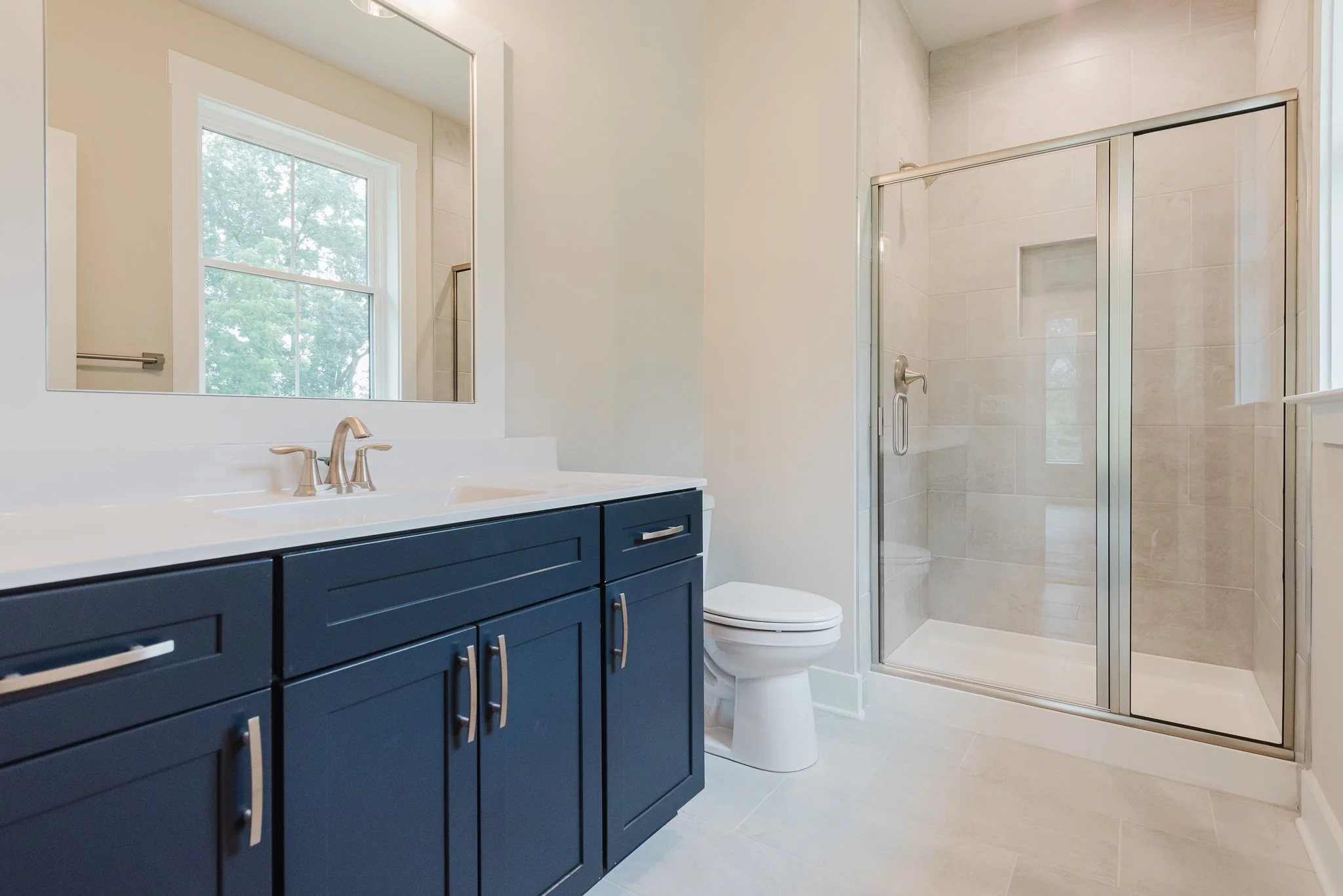 Modern bathroom with navy blue vanity, white countertop, large mirror, window, toilet, and glass shower enclosure.