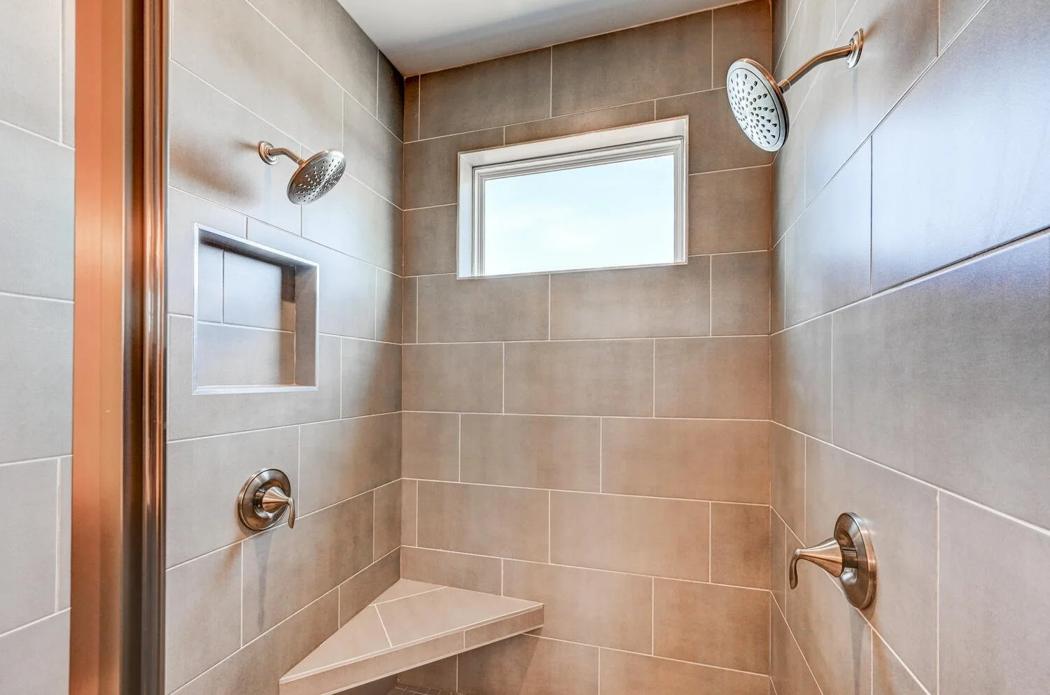 A walk-in shower with beige tiled walls, two showerheads, a built-in shelf, and a small window.