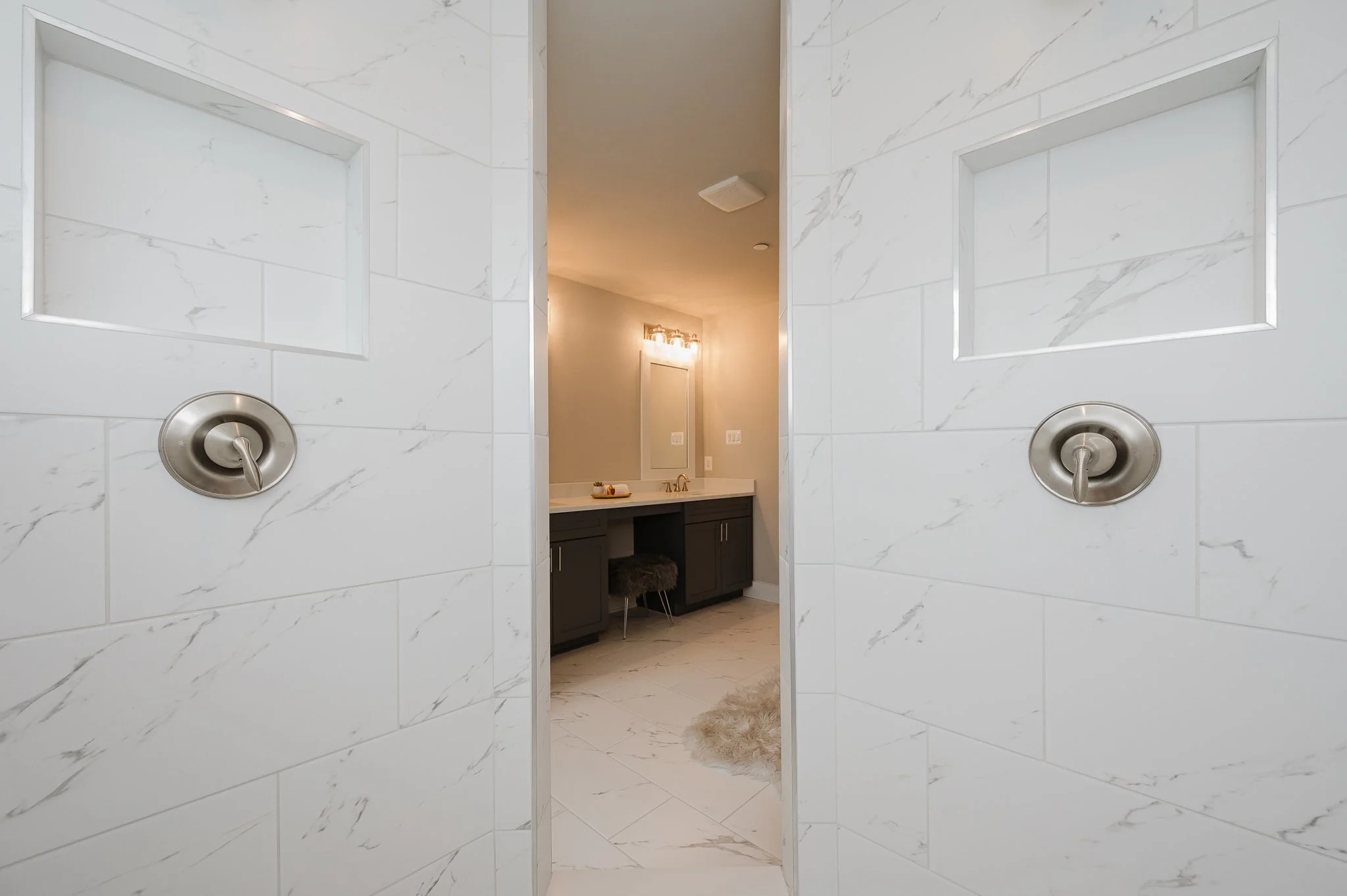 View of a bathroom shower entrance with white marble tiled walls and modern brushed nickel shower controls, leading to a bathroom vanity with drawers, a mirror, and lighting fixtures.