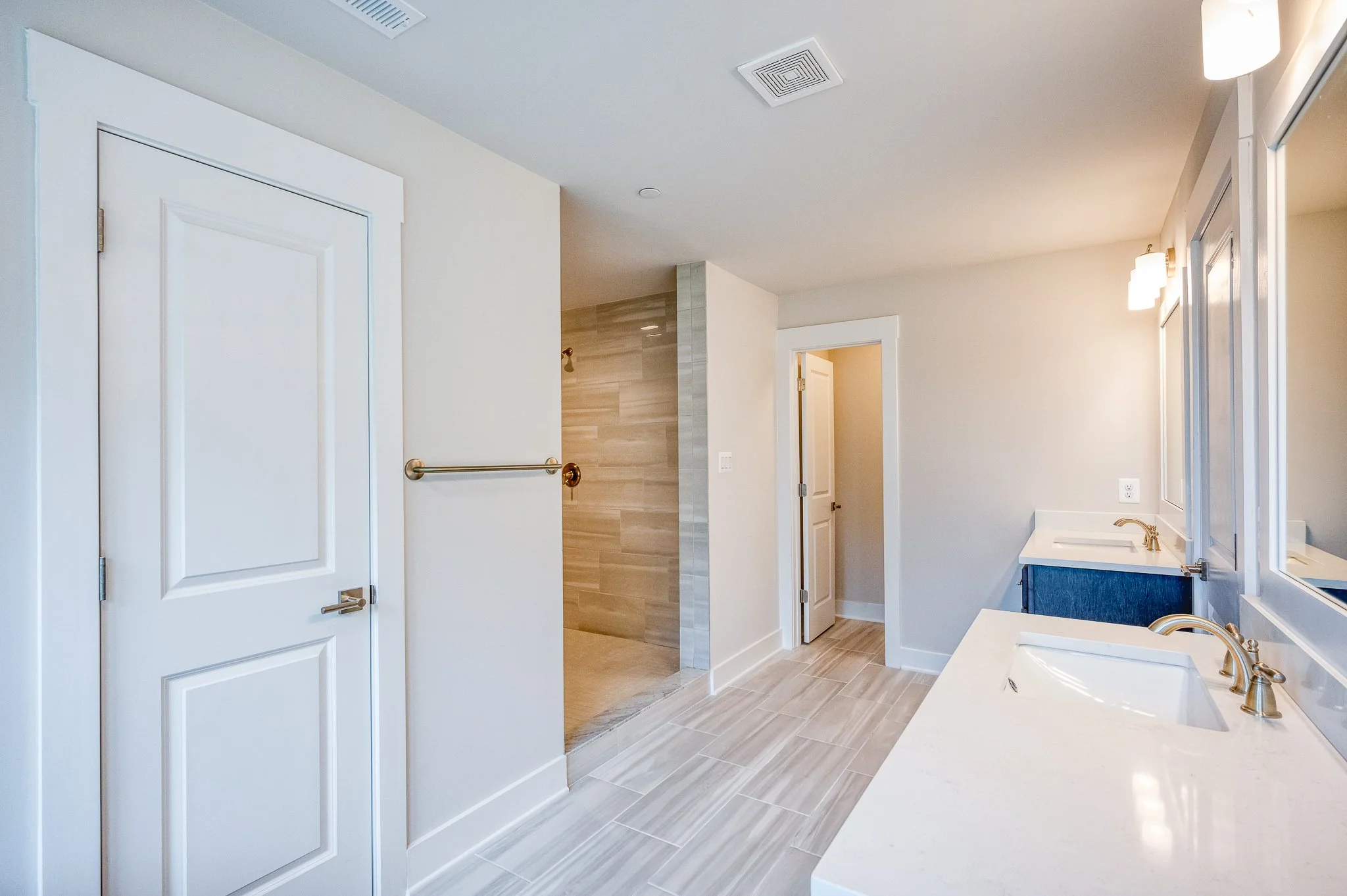 A modern bathroom with white walls, a double vanity with white countertops, gold fixtures, and large mirrors. There is a walk-in shower with beige tiles and a glass door. The bathroom has beige wood-like floor tiles, and a white door leading to anoth