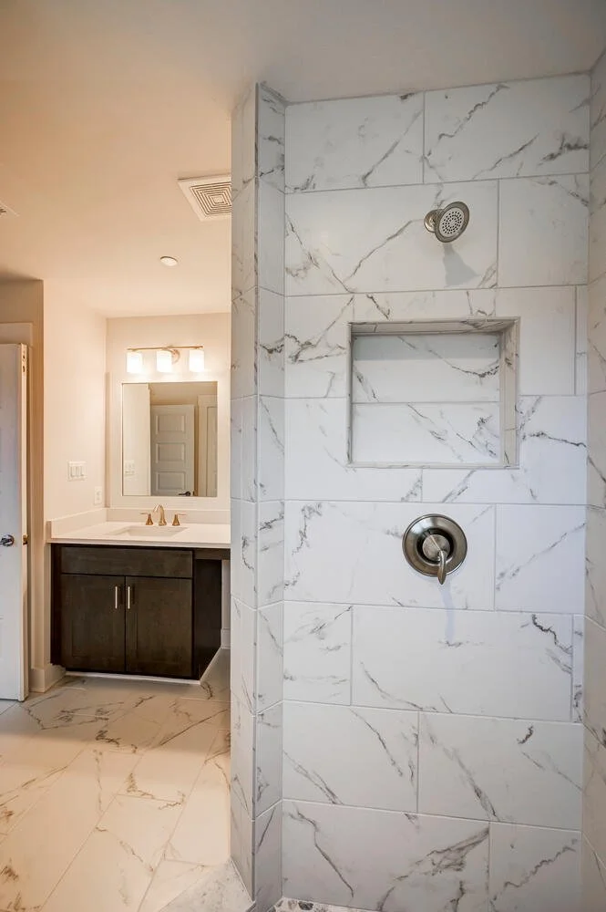 A modern bathroom shower with white marble tiles, a built-in soap niche, and a silver shower handle and head. Visible part of a dark wood vanity with a sink, faucet, and mirror in the background.