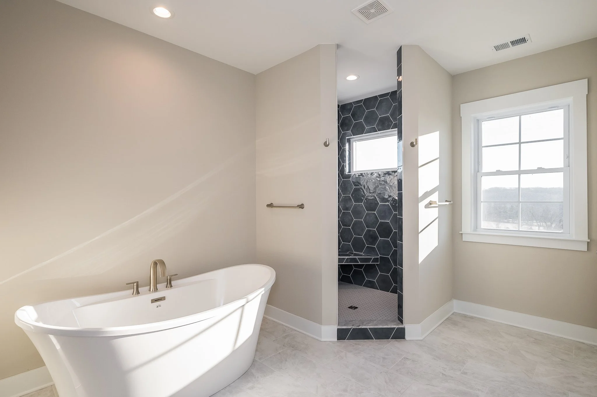 A modern bathroom featuring a white bathtub with a brushed nickel faucet, a walk-in shower with black hexagonal tiles, a small window in the shower, and a large window with a mountain view.