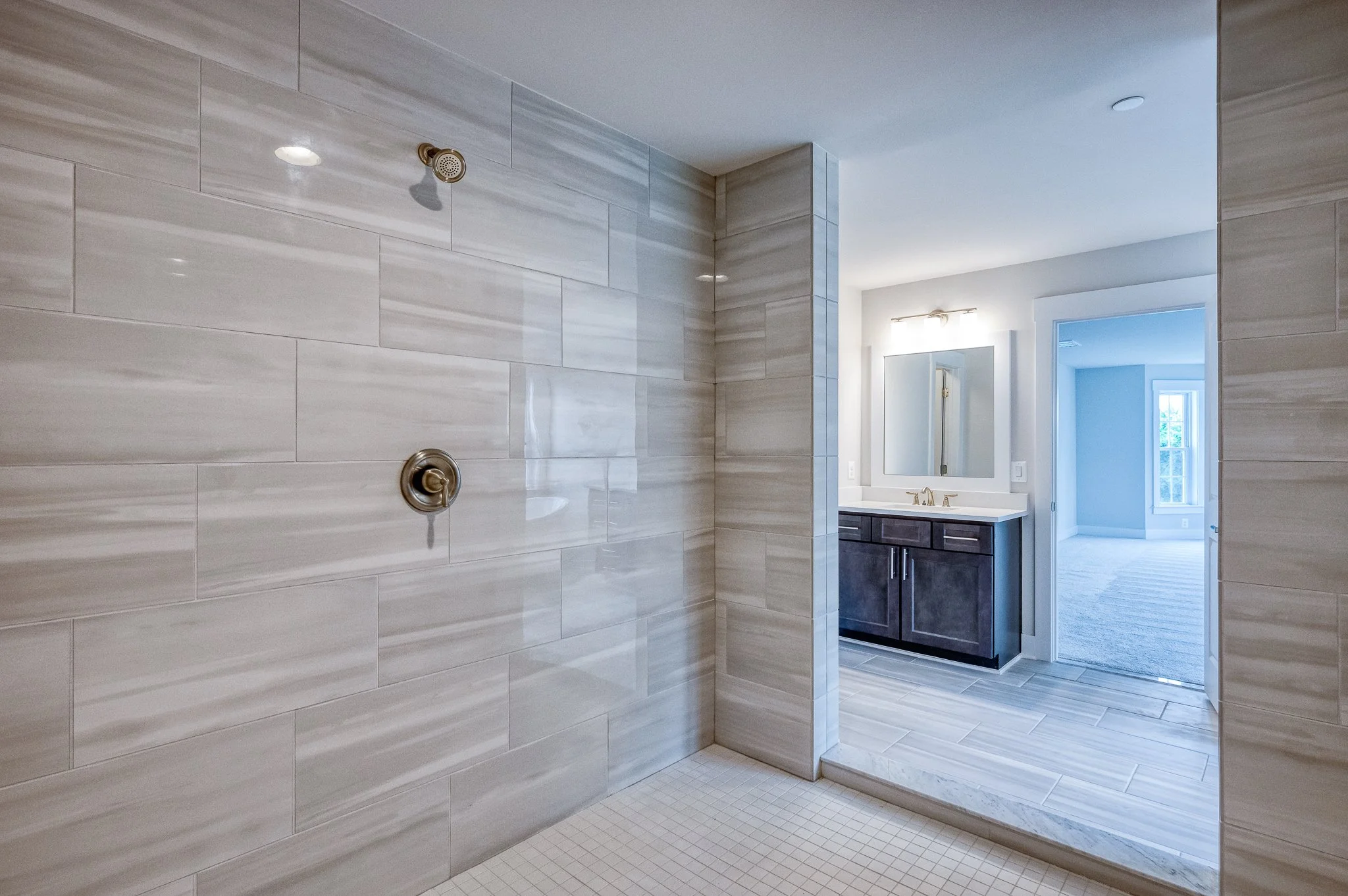 Empty walk-in shower with large beige tiles, a brass showerhead, and a separate brass control valve. Open entrance to a bathroom with a dark wooden vanity and mirror, leading to a bright room with a window.