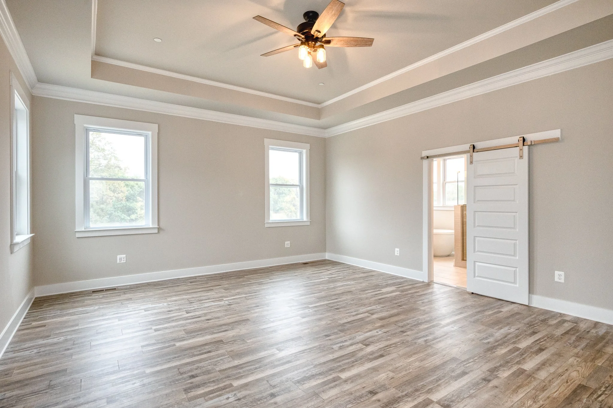 Empty living room with three windows, a ceiling fan with lights, light-colored walls, white baseboards, and a sliding barn door leading to a bathroom.