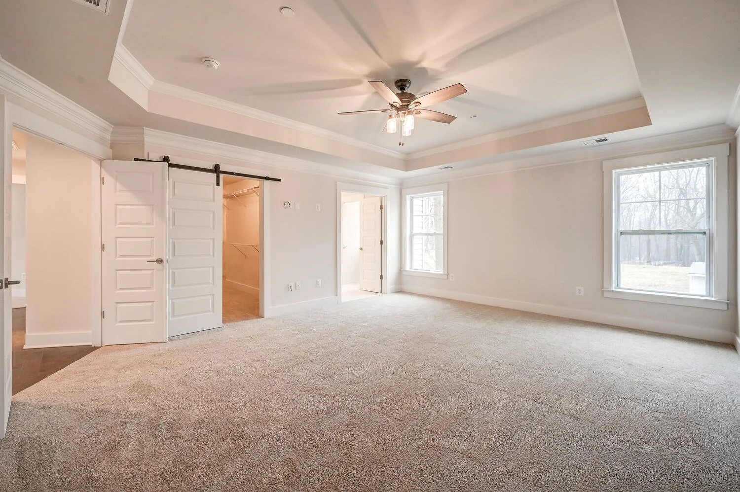 Empty living room with large windows, beige carpet, white walls, and a ceiling fan.