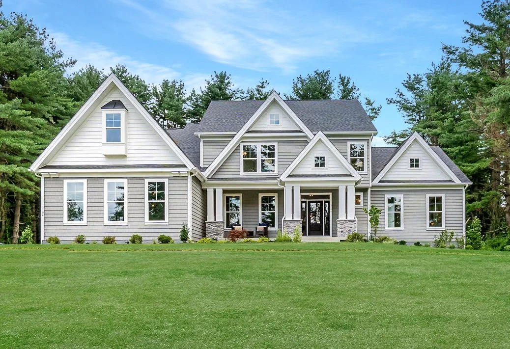 Large modern house with gray siding, multiple gable roofs, and a front porch set against a green lawn and trees.