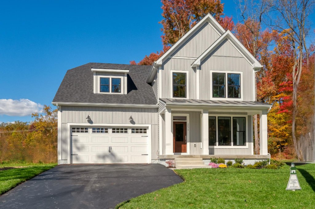 A two-story modern house with white vertical siding, a gray shingled roof, a front porch with white columns, and a landscaped lawn with green grass and colorful bushes, set against a background of trees with autumn foliage and a clear blue sky.