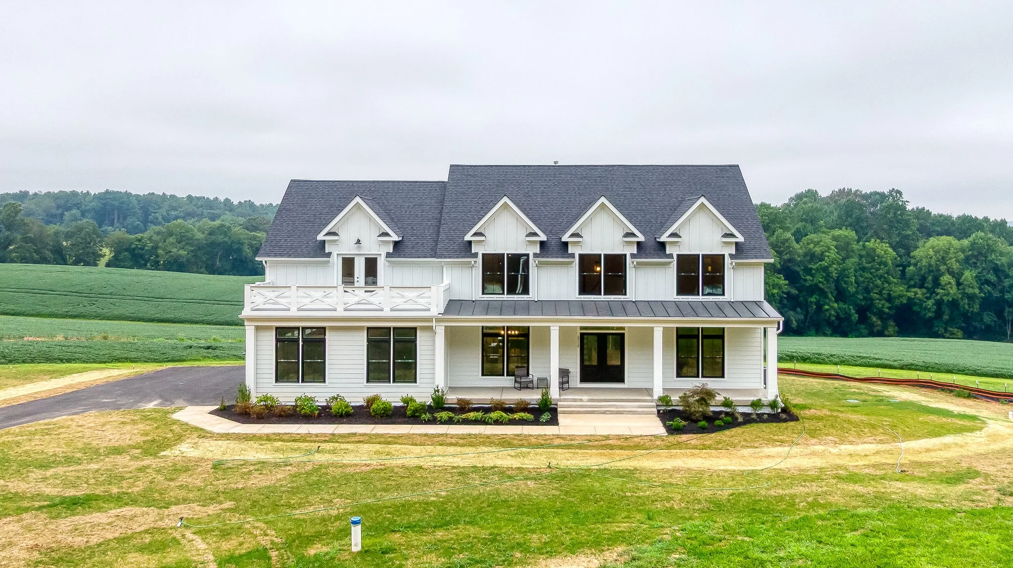 A modern two-story white house with black roof, surrounded by green fields and trees, with a small front porch, black window frames, and a landscaped front yard.