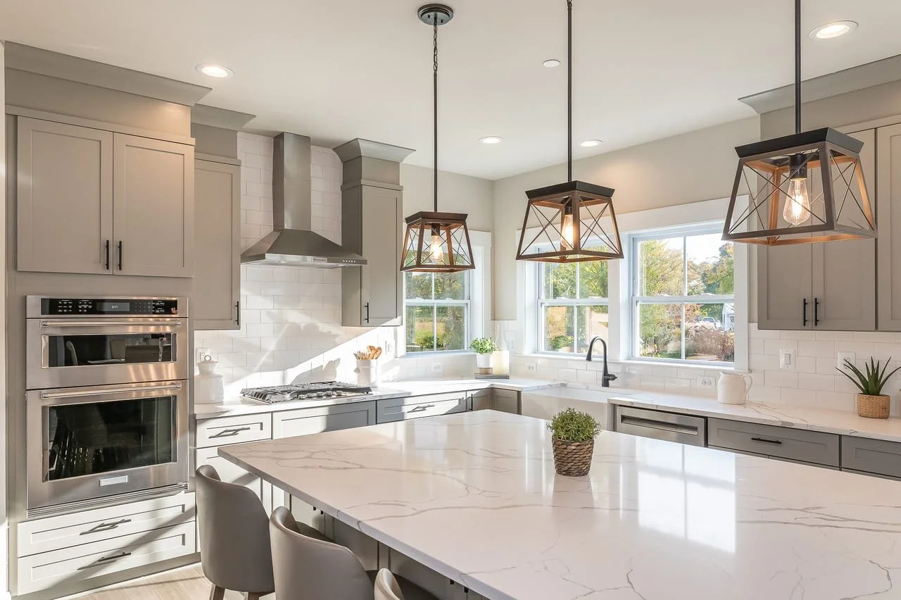 Modern kitchen with gray cabinets, white marble countertops, a central island, stainless steel appliances, pendant lights, and large windows with a view of trees.