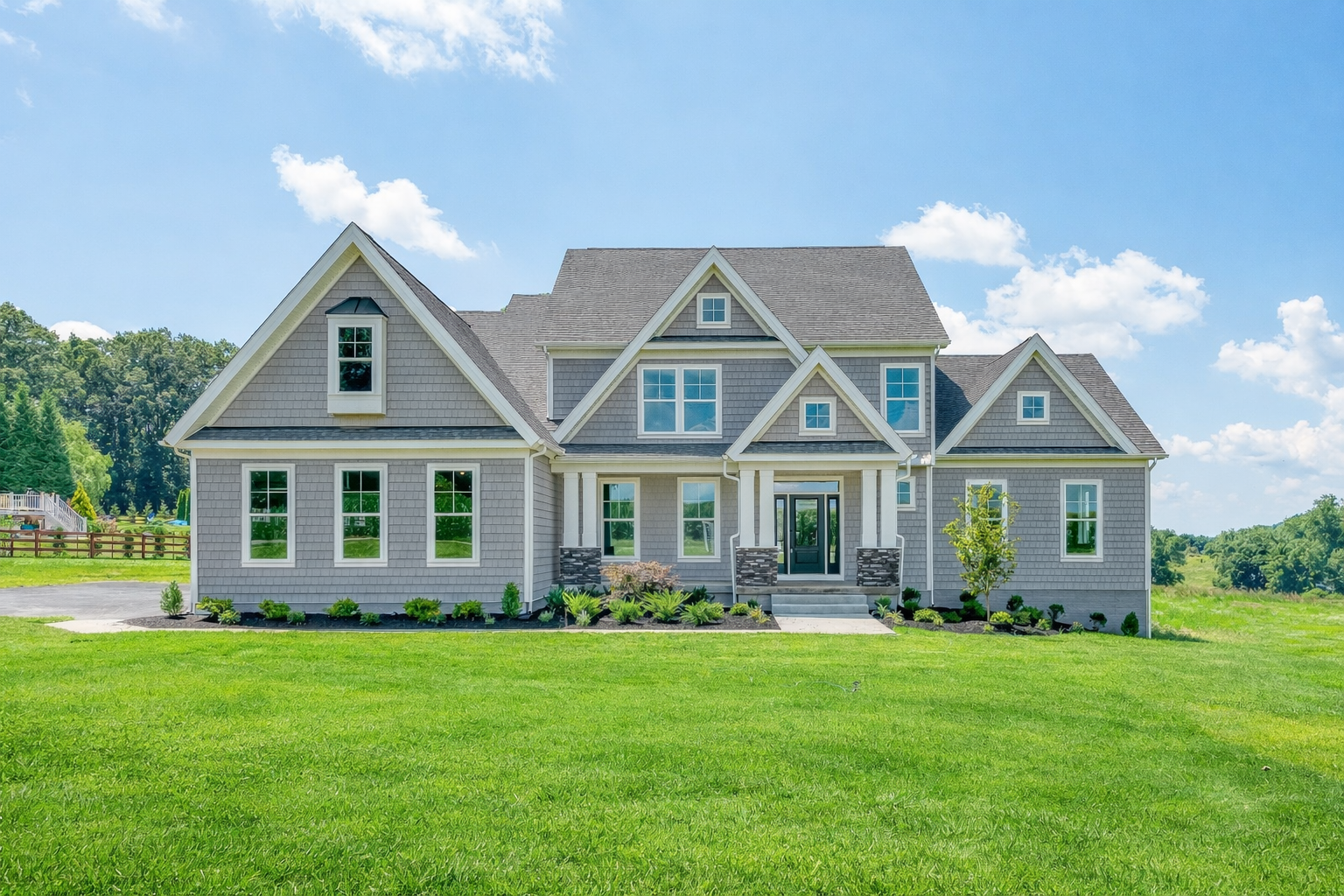 A large, modern house with a gray exterior and multiple roof gables. It is surrounded by a green lawn and landscaping, with a bright blue sky and scattered clouds in the background.