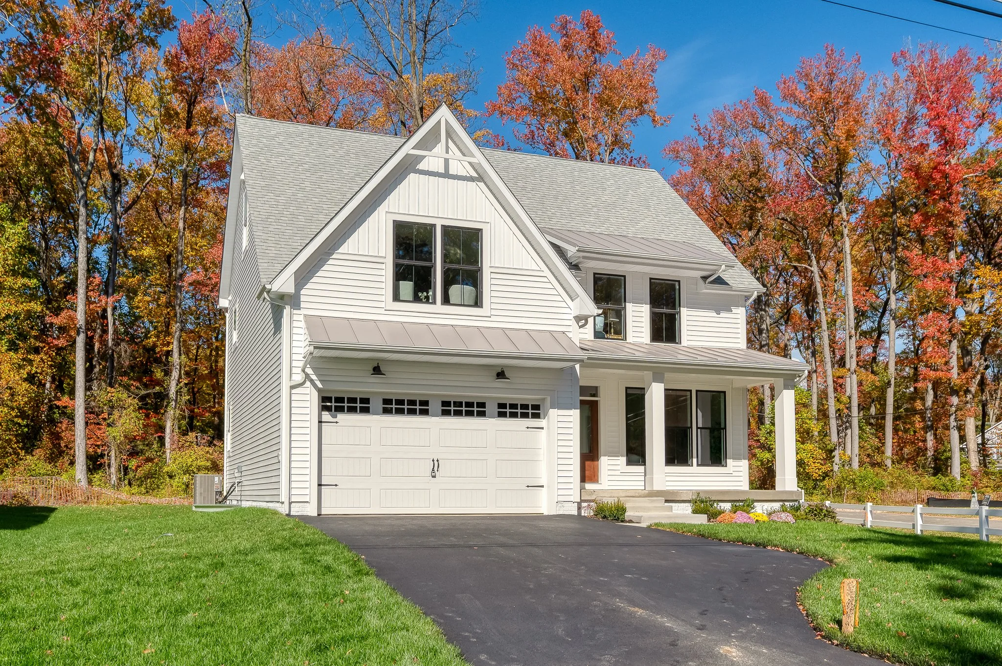 White two-story house with a garage, surrounded by green lawn and trees with autumn foliage, under a clear blue sky.