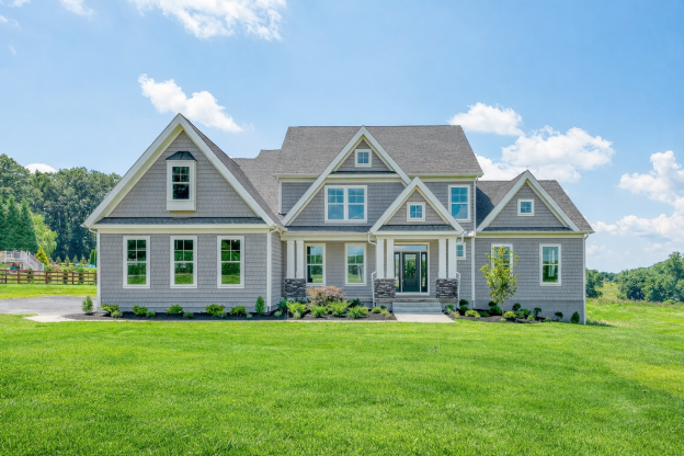 Modern two-story house with gray siding, multiple gables, and front steps, set on a grassy lawn with trees in the background on a bright, sunny day.