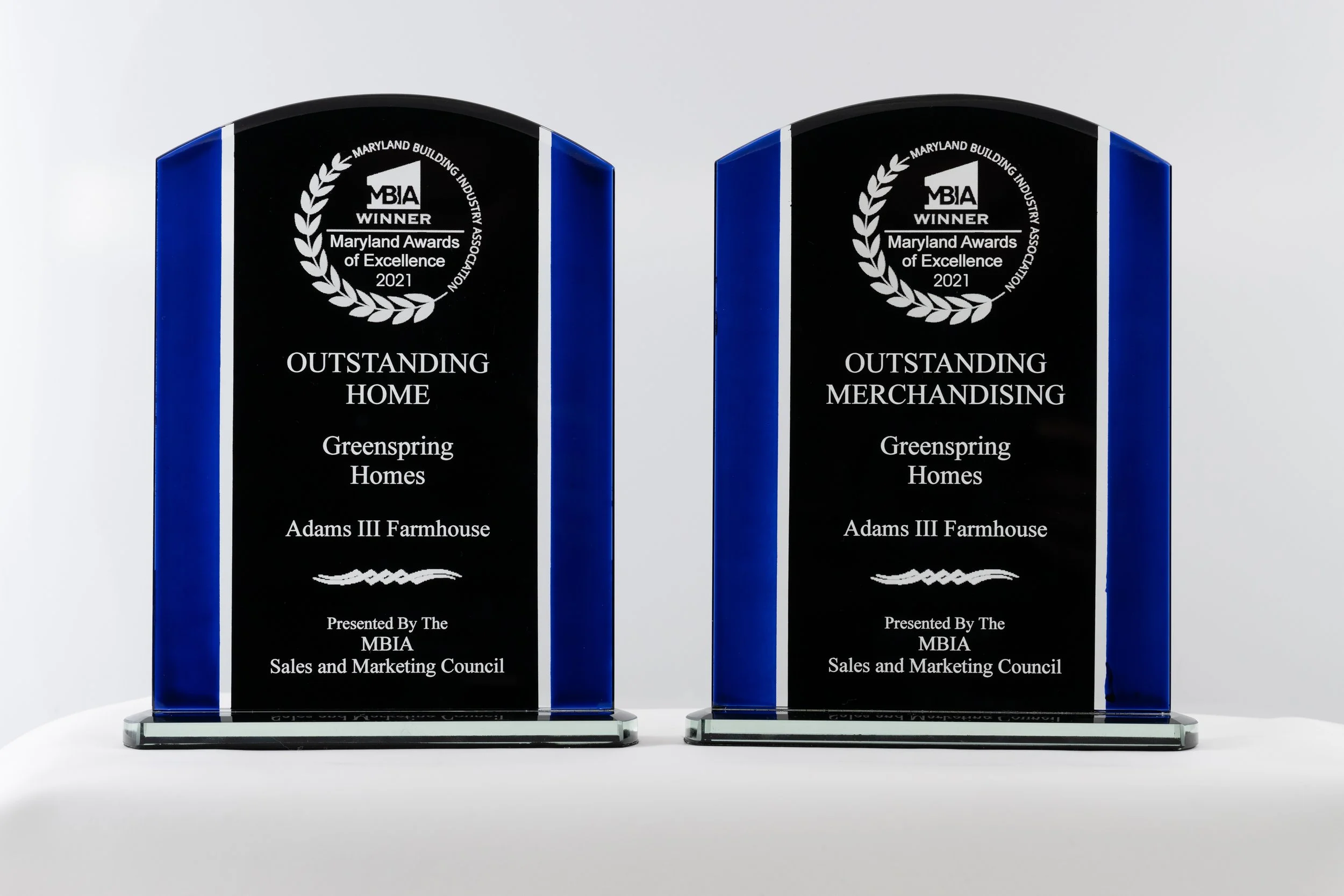 Two glass awards with blue edges, each inscribed with the Maryland Building Industry Association award for 2021. The left award is for 'Outstanding Home' and the right for 'Outstanding Merchandising,' both presented to Greenspring Homes for Adams III Farmhouse, by the MBIA Sales and Marketing Council.