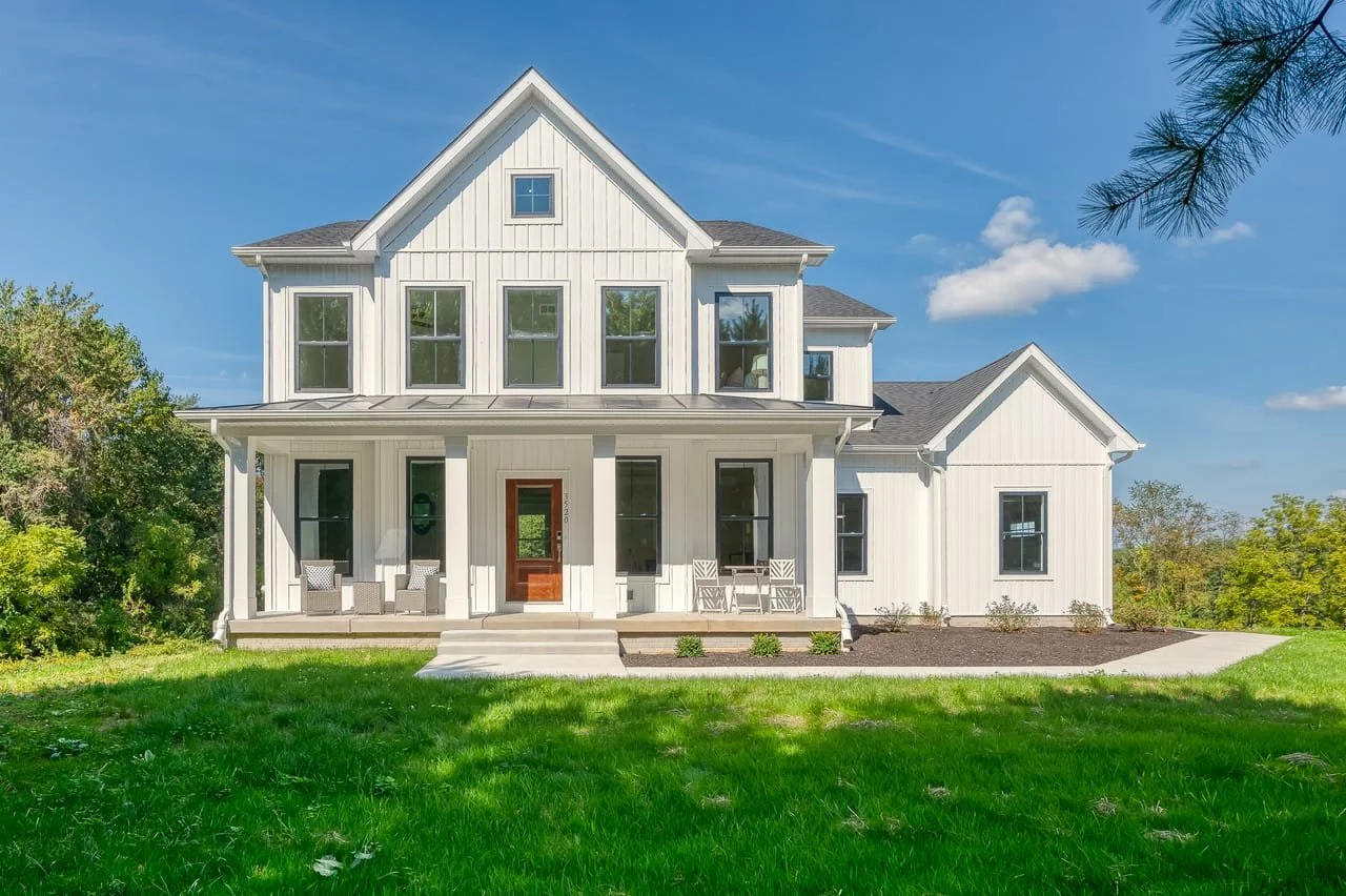 A two-story white house with a front porch, surrounded by green grass, trees, and a clear blue sky.
