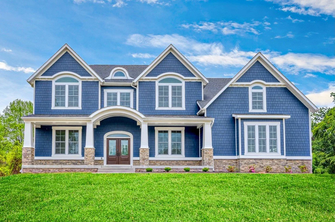 A two-story dark blue house with white trim, multiple gabled roofs, and large windows, surrounded by green trees and lawn.