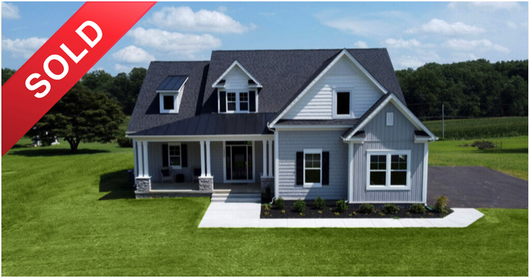 A newly sold two-story house with a front porch, white siding, and a dark roof, surrounded by a green lawn and a paved driveway.