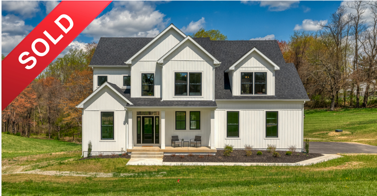 A newly built white two-story house with black roof, large front porch, and multiple windows, located in a green grassy area with trees in the background, marked with a red 'SOLD' banner.
