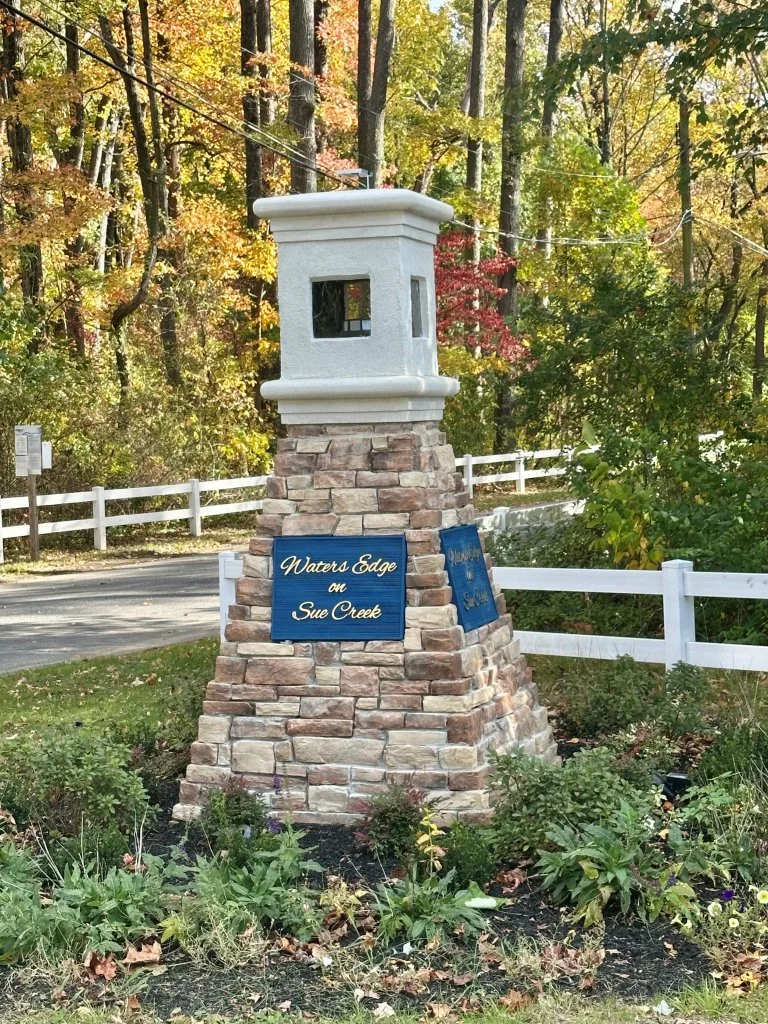 A wooden and brick lighthouse-shaped sign with two blue plaques that read "Waters Edge on Sue Creek," situated in a garden with autumn-colored trees and a white fence in the background.
