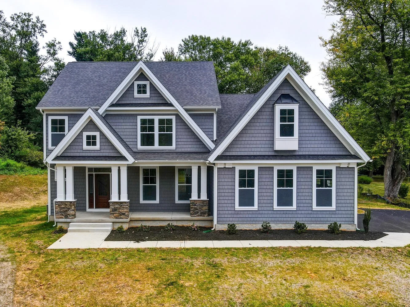 A modern two-story gray house with white trims, multiple gables, and large windows, situated in a grassy yard with trees in the background.