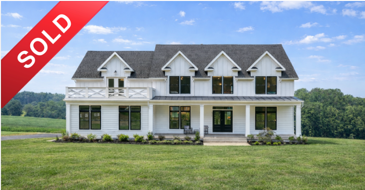 Large white two-story house with dormer windows, a balcony, and a front porch, located in a grassy field with trees in the background, marked as sold.