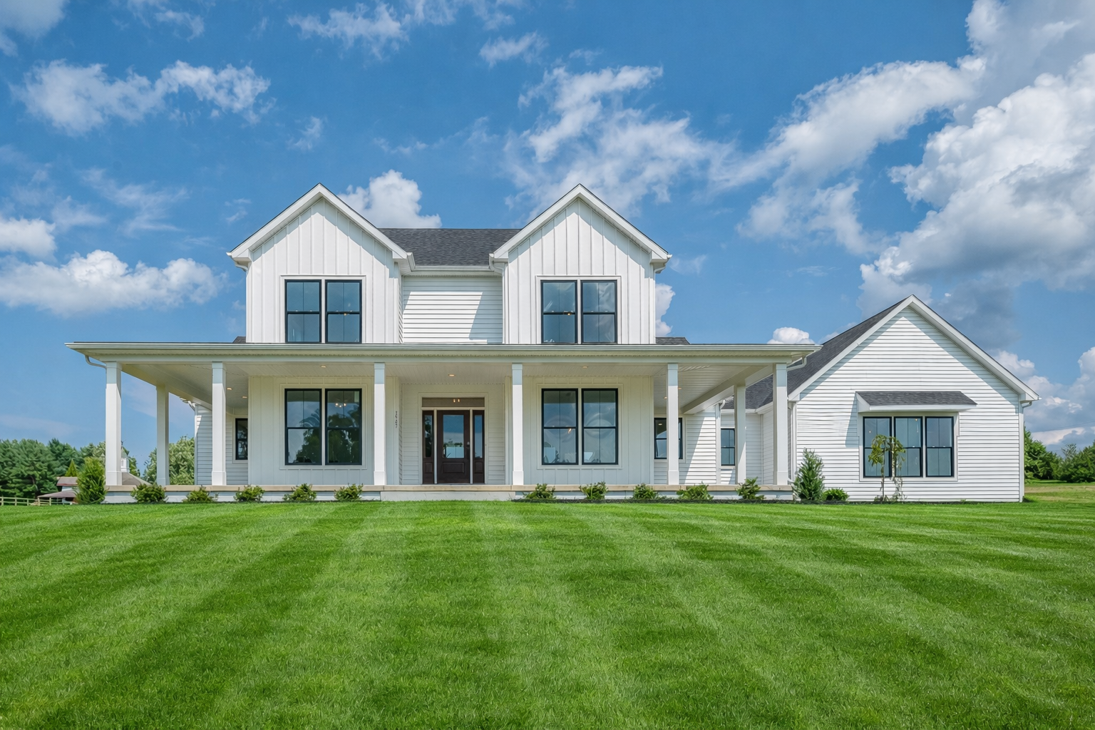 A large modern house with gray and white siding, multiple gabled roofs, and a front porch with furniture, situated on a grassy lawn with a black barrier in the foreground.