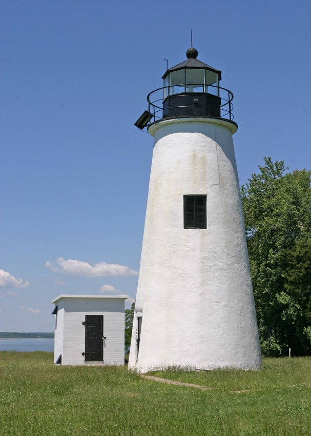 A white lighthouse with a black lantern room and railing, beside a small white building, set against a clear blue sky and green trees.