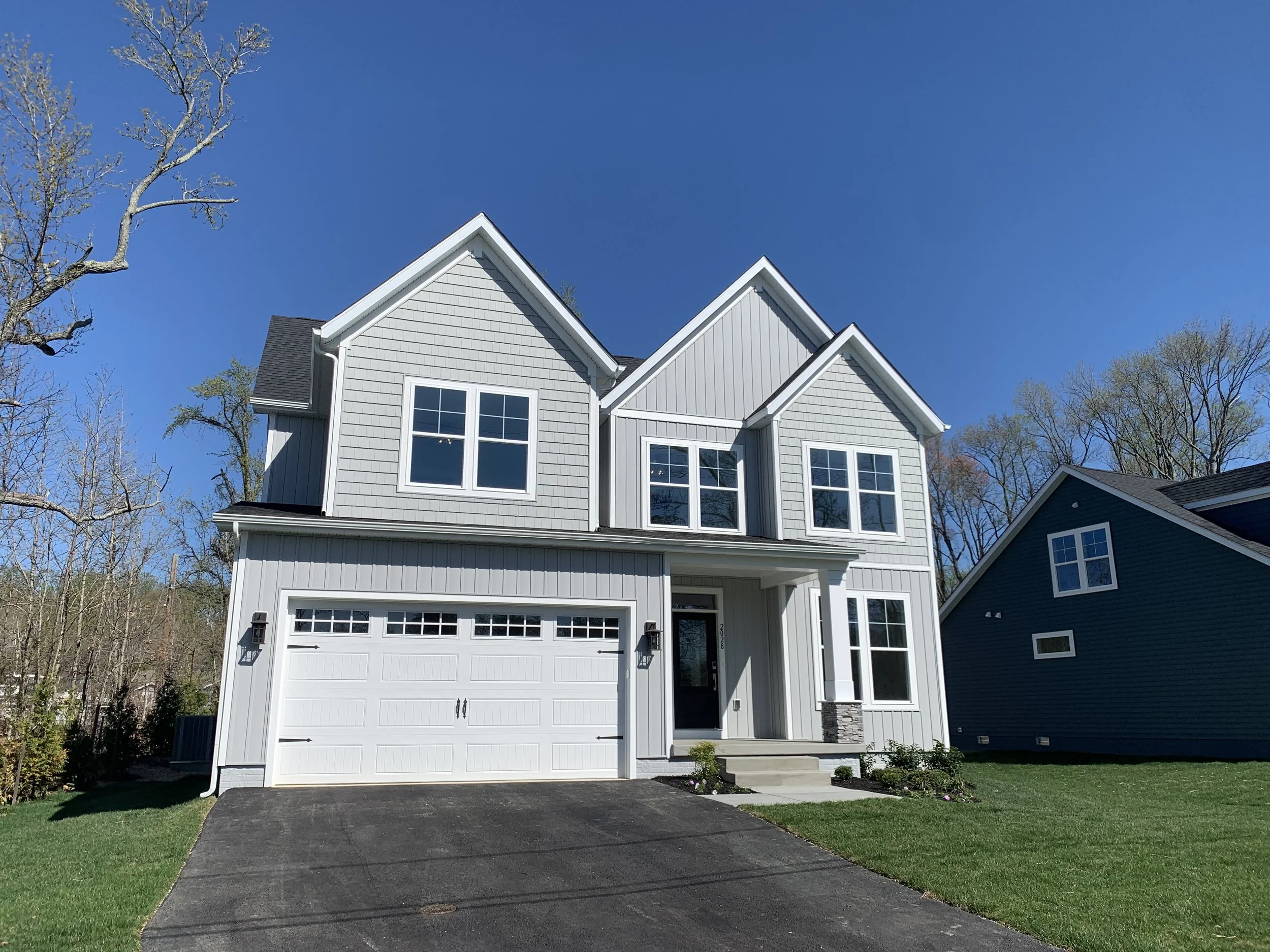 A two-story house with blue siding, white trim, and a front porch with a wooden door. The house has a two-car garage on the right and is surrounded by green trees and shrubs under a partly cloudy sky.