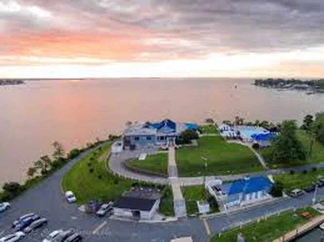 Aerial view of a lakeside property with a large house, shorter structures, parking lot, and green lawns at sunset.
