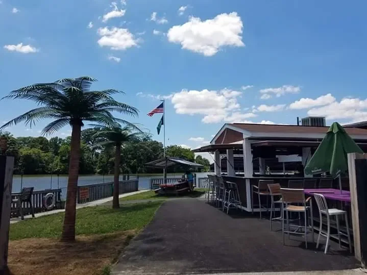 Outdoor scene with two artificial palm trees, a boat docked near a house, and a patio area with high chairs and tables under a green umbrella. The sky is partly cloudy.
