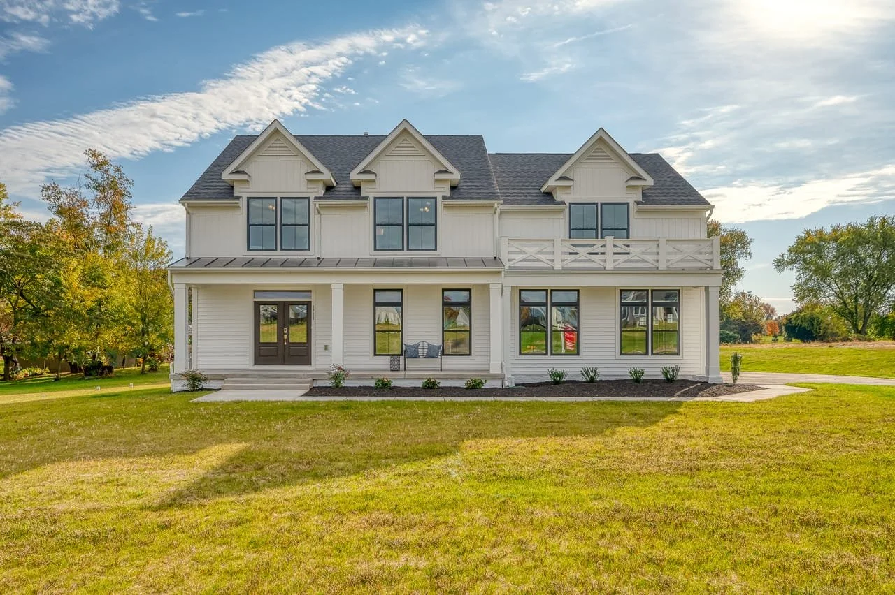 A large white, multi-story house with a gray roof, multiple windows, a small front porch, and a balcony in a grassy yard with trees in the background.