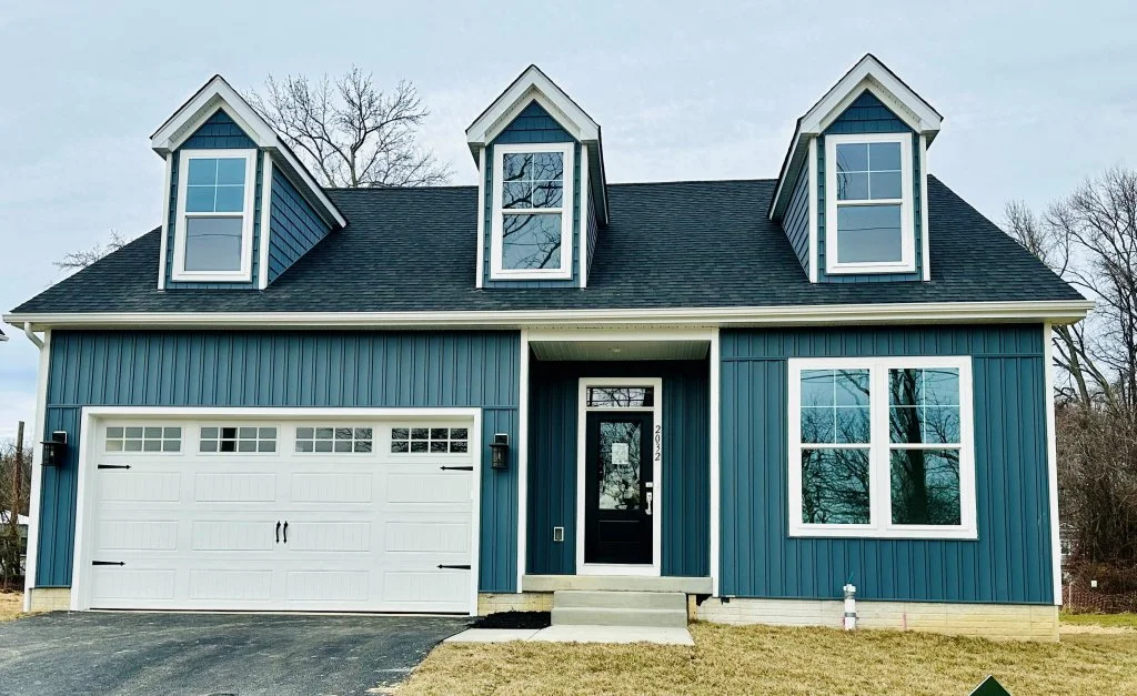 New blue suburban house with dormer windows, a white garage door, and a black front door, surrounded by a lawn and leafless trees.