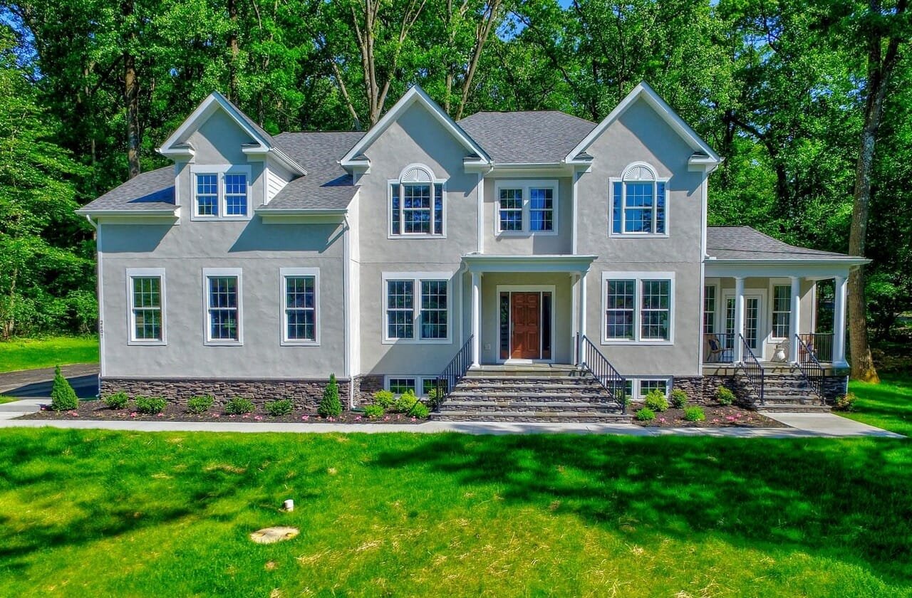 A large, two-story house with a gray exterior and a dark gray roof, surrounded by green trees and lawn, with steps leading to the front door and a porch on the right side.