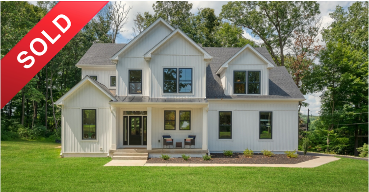 A newly built white two-story house with a landscaped front yard and a concrete walkway. The house has multiple windows, a front porch with chairs, and steep gabled roof sections. A red sold banner overlays the top left corner.