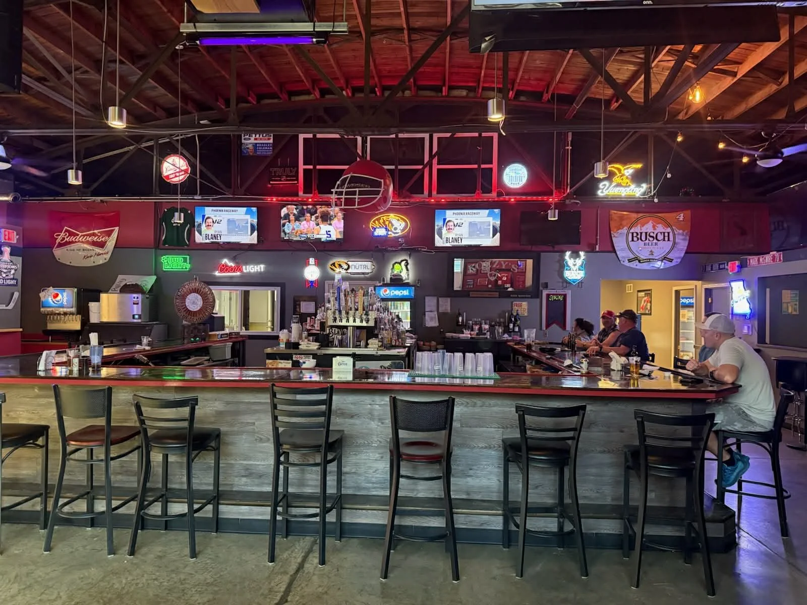 Interior of a bar with a curved counter, several patrons seated, neon signs, multiple television screens, and sports event advertisements.