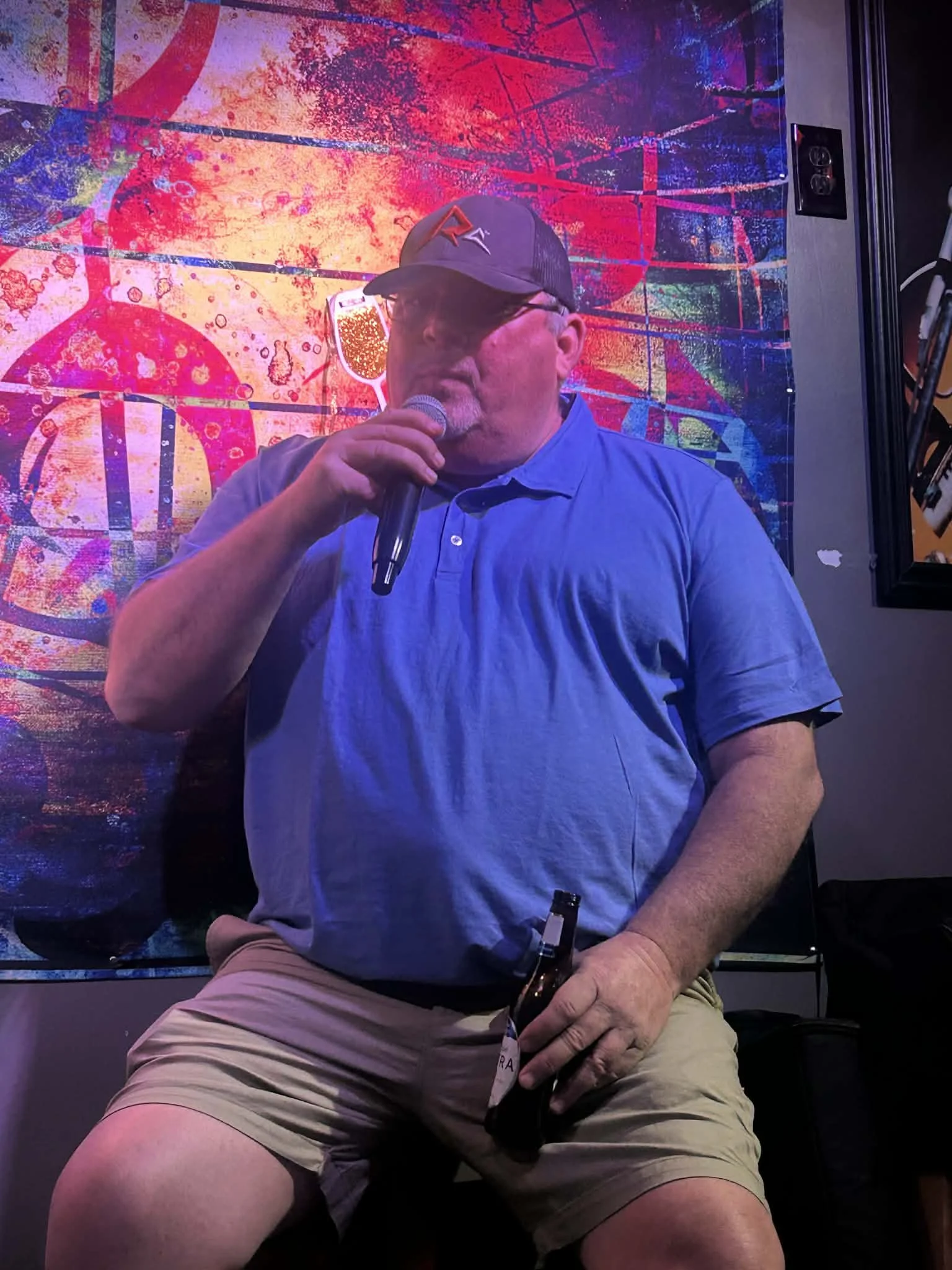 A man in a blue polo shirt and beige shorts holding a microphone and a beer bottle, sitting in front of a colorful abstract neon artwork.