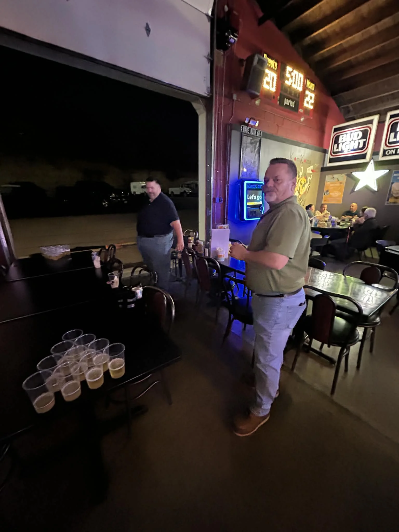 Two men standing inside a bar near a large open garage door at night. One man is wearing a green shirt and khaki pants, holding a drink, while the other is in a dark shirt and jeans."