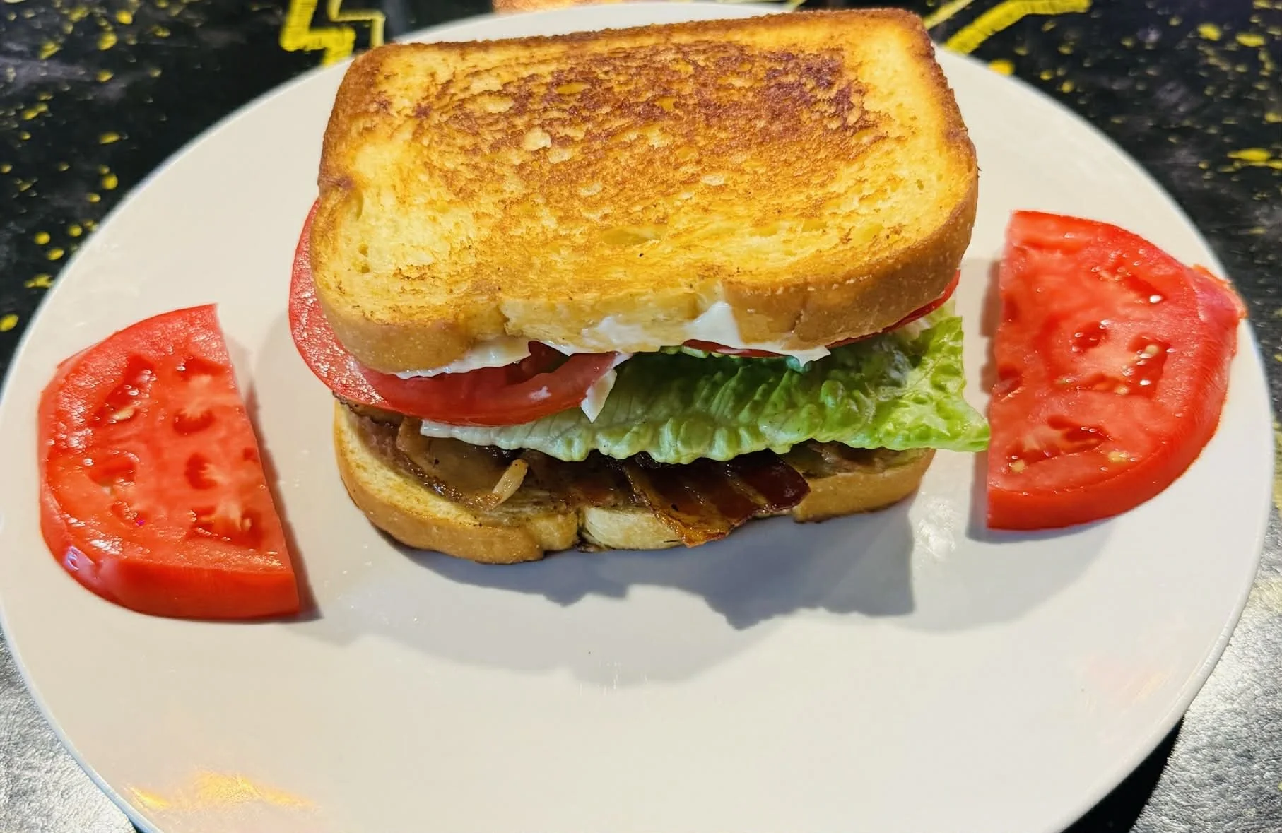 A close-up of a bacon cheeseburger with lettuce, tomato, and a toasted bun, served with two slices of tomato on a white plate.