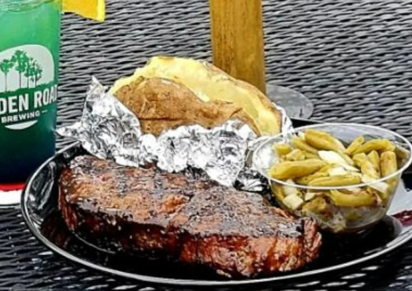 A plate of barbeque ribs, a baked potato with toppings, and a side of green beans, with a green can of Golden Road Brewing behind the plate.