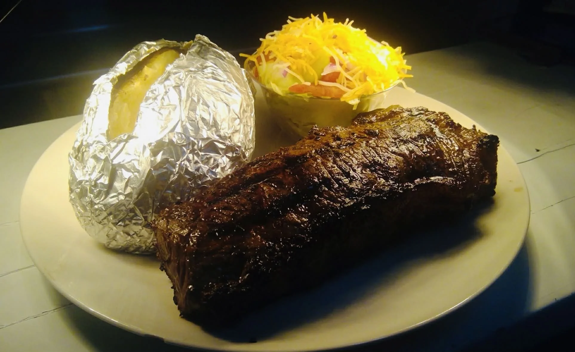 Plate with baked potato, grilled beef short ribs, and a side of shredded cheese salad in a small bowl.