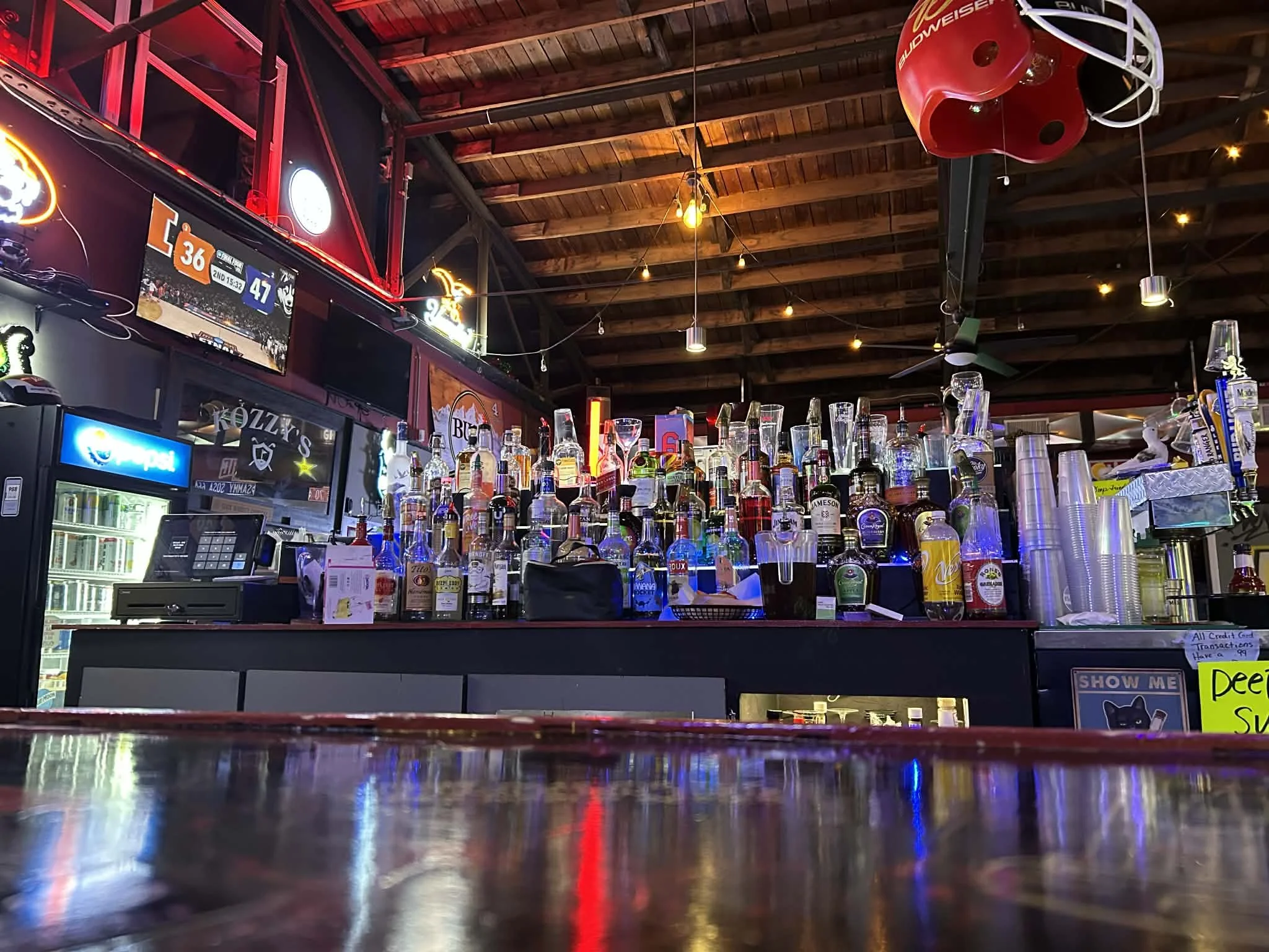Bar interior with liquor bottles on shelves, neon signs, television screens, and a wooden ceiling.