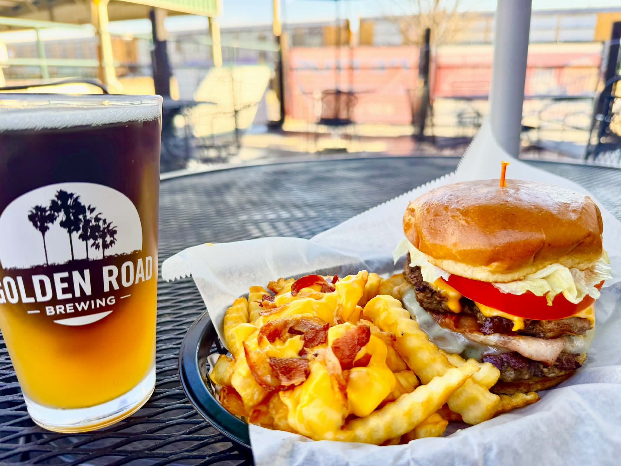 A burger with lettuce, tomato, cheese, bacon, and multiple beef patties, with a toothpick on top, served with French fries and cheese, alongside a pint of beer with the Golden Road Brewing logo, on a black metal outdoor table on a sunny day.