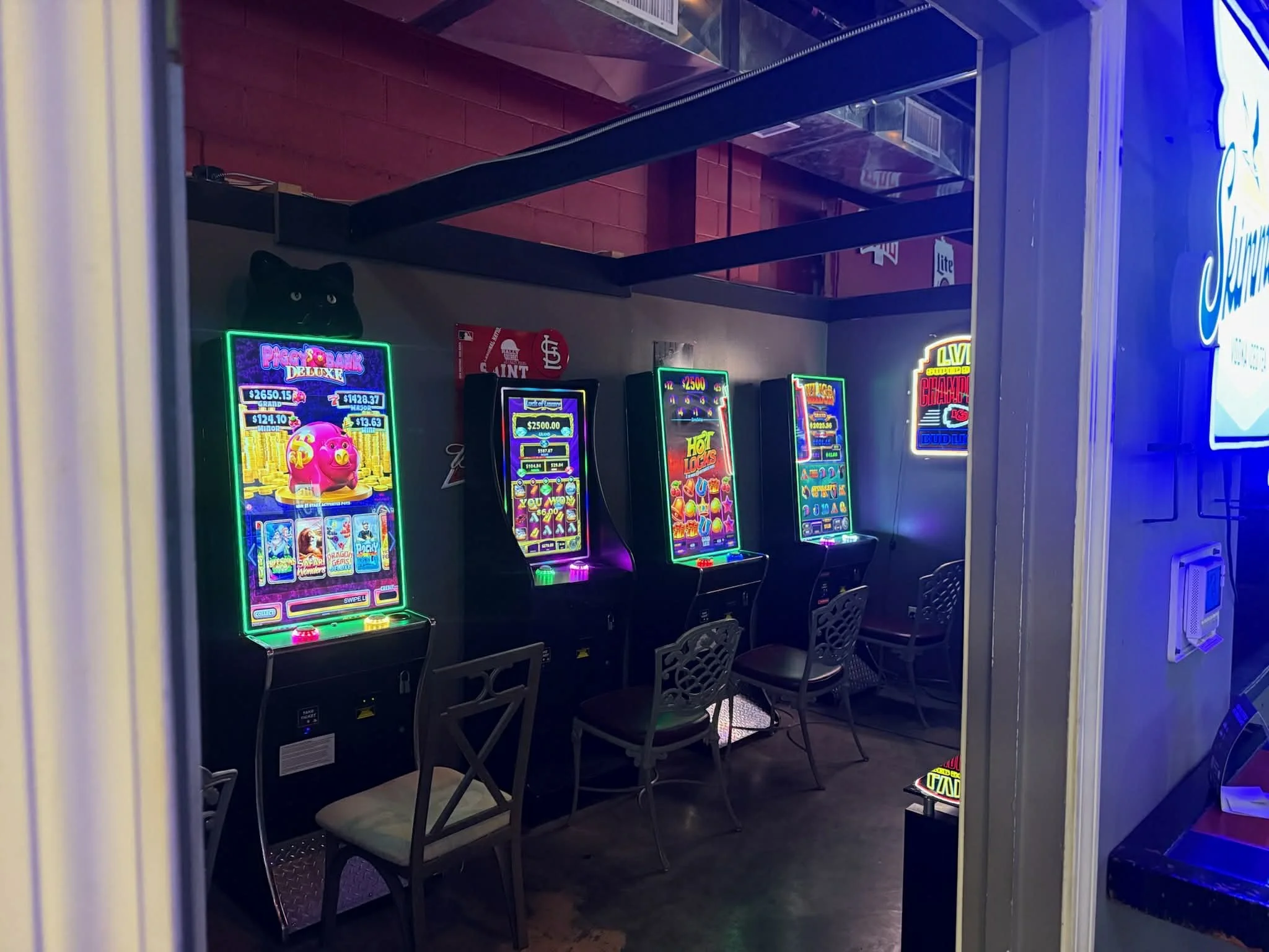 A row of four brightly lit slot machines inside a gaming room with chairs in front of each machine. The machines display colorful graphics and symbols, and there is a neon sign on the wall behind them.