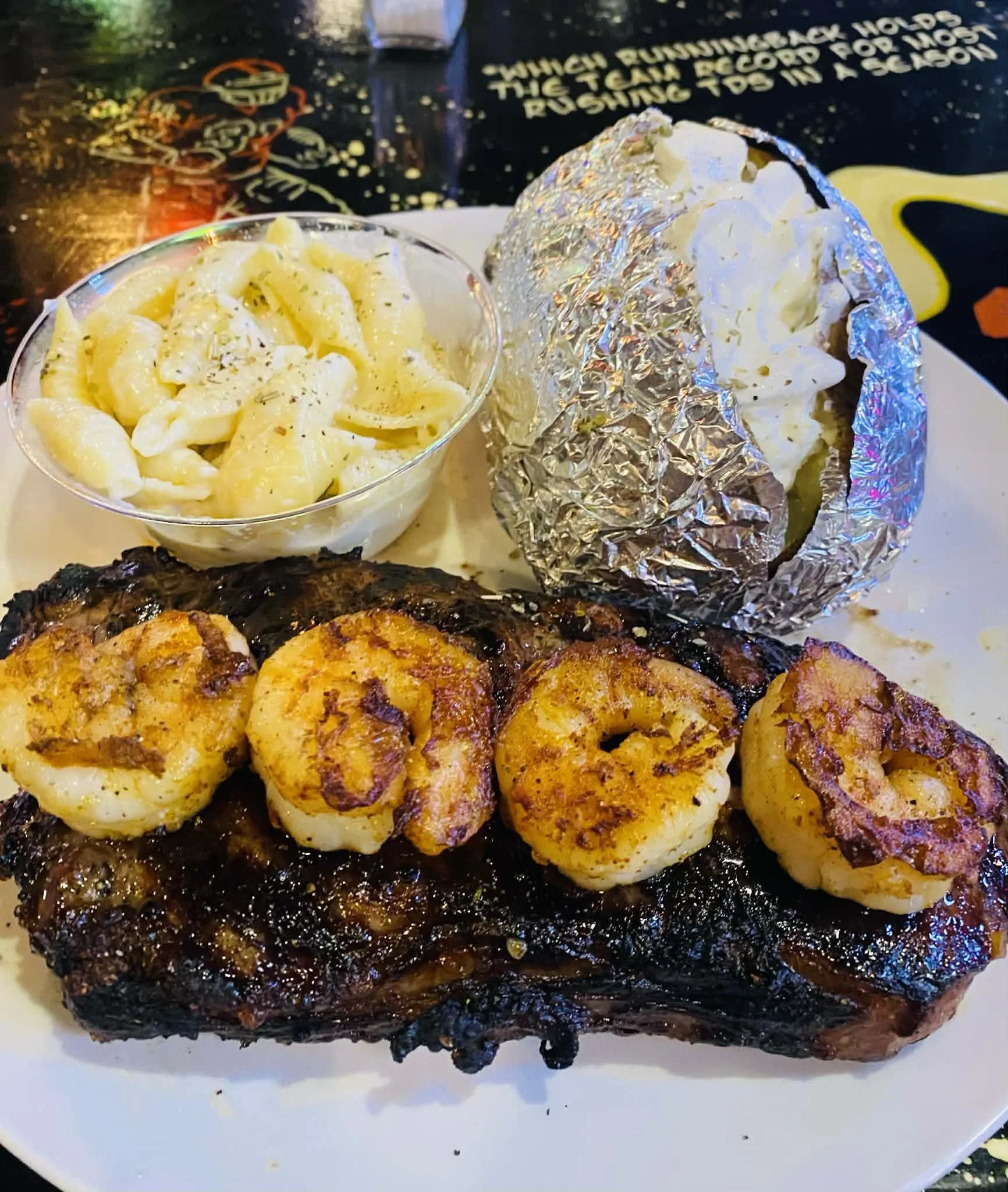 Steak topped with grilled shrimp, served with macaroni and cheese, and a baked potato wrapped in aluminum foil.