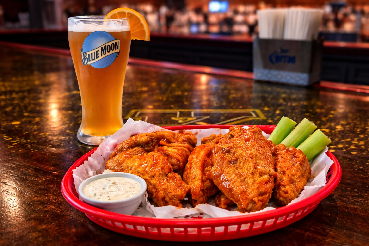 A glass of beer with an orange slice, a basket of fried chicken wings with celery sticks and ranch dipping sauce on a bar counter.