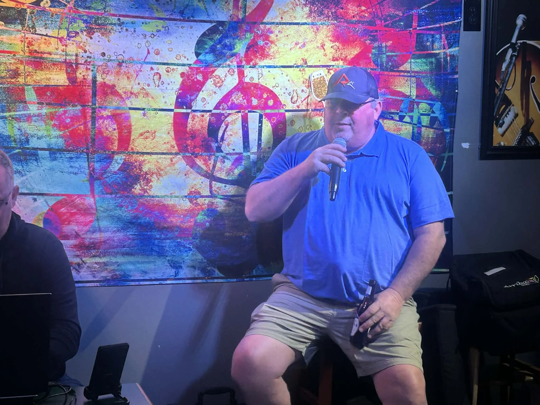 A man wearing a blue shirt, beige shorts, and a baseball cap is sitting and speaking into a microphone. Behind him is a colorful, abstract wall with music notes and symbols.