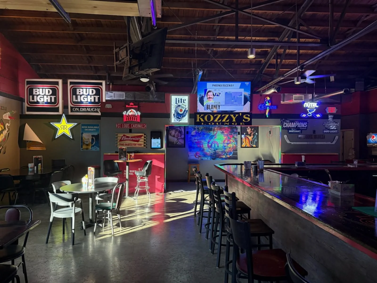 Interior of a sports bar with tables, chairs, neon signs, and multiple TV screens on red painted walls, illuminated by natural light from windows.