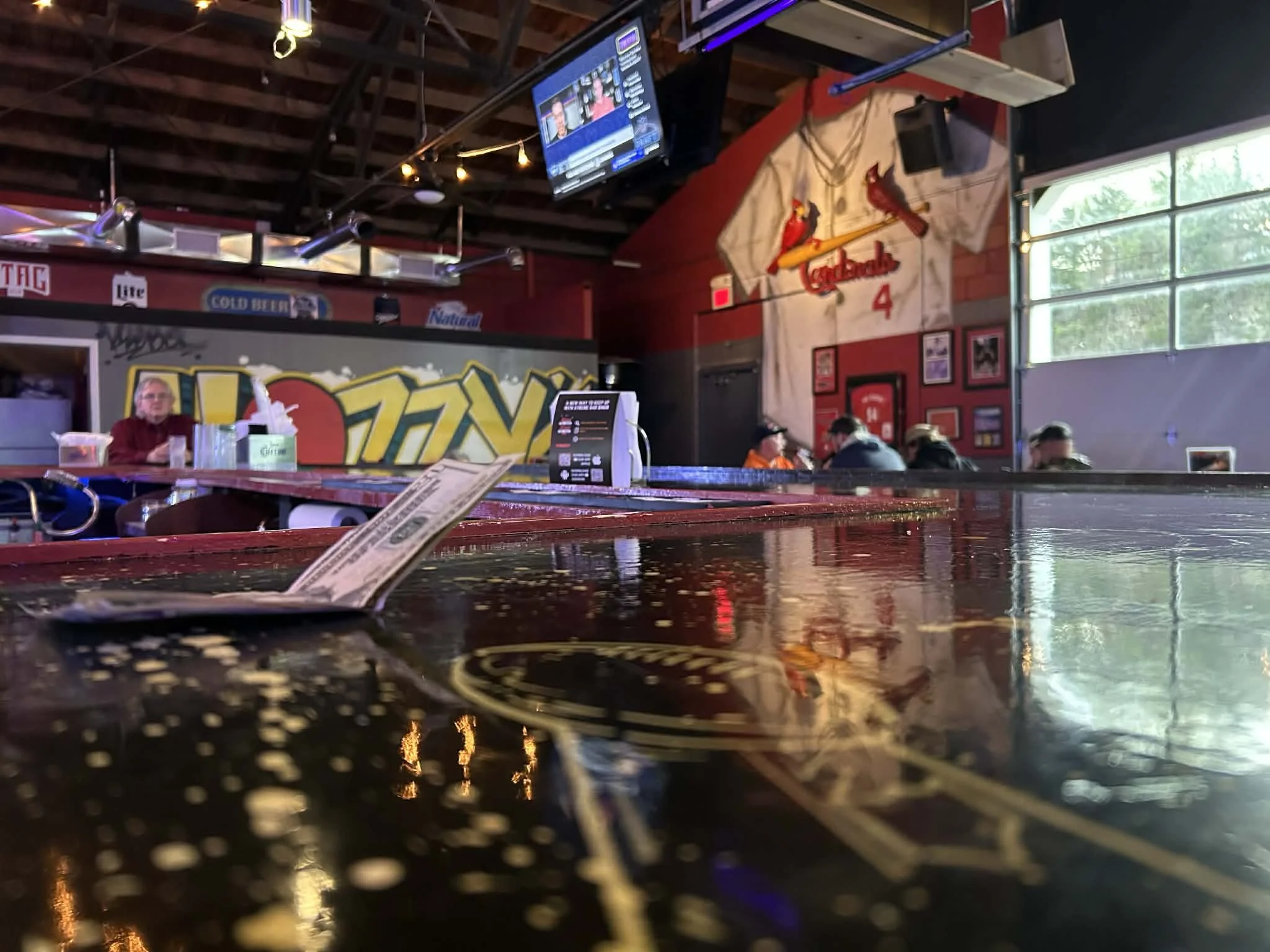 Interior of a casual restaurant or bar with a mural of the St. Louis Cardinals logo of a cardinal bird on the wall. A jersey with the number 4 hangs on the wall, and there are patrons seated at tables. A TV is mounted on the ceiling, and the foreground shows a glossy table with a receipt or menu. The setting has a cozy, sporty atmosphere with beer advertisements and framed photos on the walls.