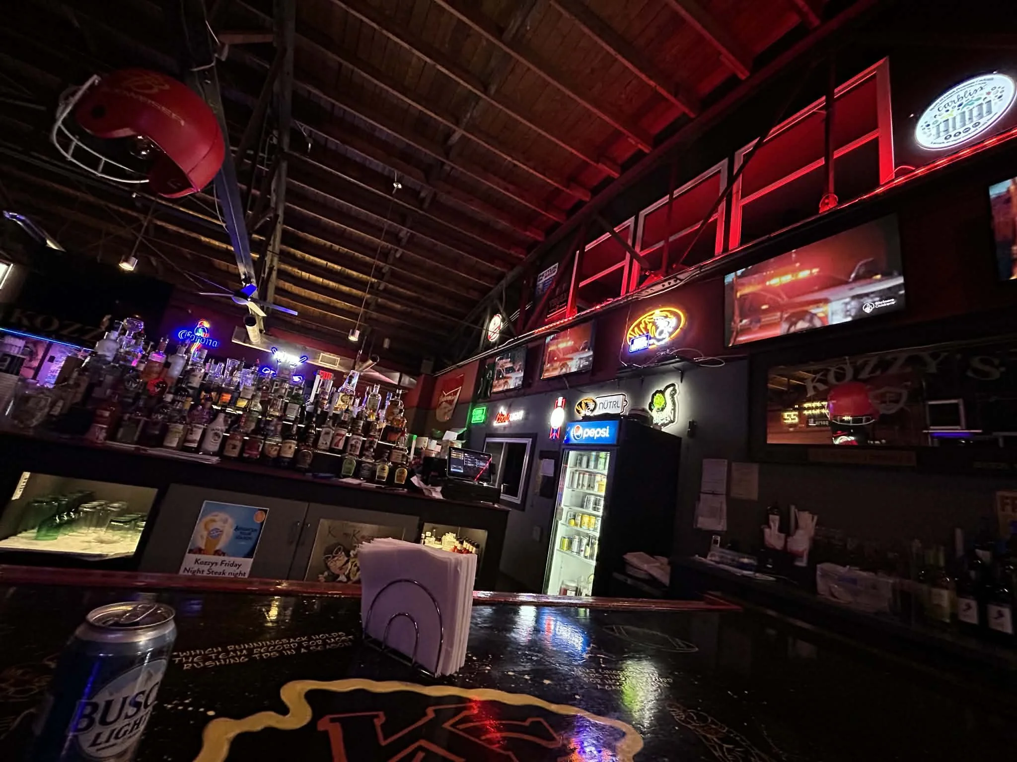 A dimly lit bar with a variety of liquor bottles on shelves behind the counter. Neon signs and advertisements, including Pepsi and Corona logos, are visible on the walls. There is a refrigerator with drinks, a bar stool, and a beer helmet hanging from the ceiling.